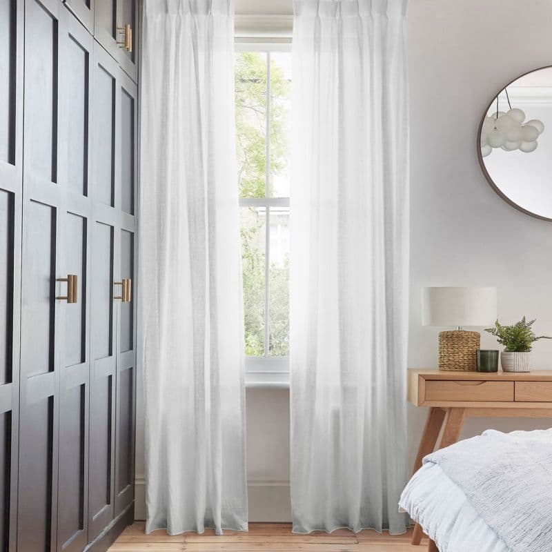 Sheer white pleated curtains, floor-length and hung on either side of a tall sash window, filtering soft daylight; bedroom setting with dark paneled wardrobe, wooden bedside table, lamp and mirror.