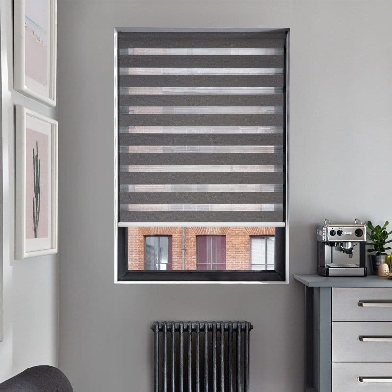 Grey zebra roller blind, lowered with alternating opaque and sheer horizontal stripes, filtering light and revealing a bottom view of a brick building; kitchen corner with radiator and espresso machine.