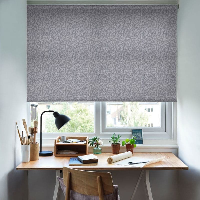 Patterned roller blind (grey leaf motif) lowered across the upper window, filtering daylight; home workspace beneath with wooden desk, lamp, potted plants and art supplies against neutral walls.