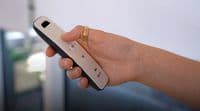 Slim white-and-black remote control held in a right hand, thumb pressing a circular my button; indoor, softly lit room with a blurred window or glass door in the background.
Text: my V somfy