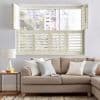 Cream louvered plantation shutters across lower sash, top hinged shutters folded outward; louvers partially open filtering daylight, city view; beige sofa with pillows, side table and lamp in living room.