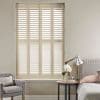 Cream plantation shutters fill a recessed window, full-height panels with horizontal louvers angled to admit light, filtering bright daylight into a neutral bedroom with armchair, bedside table and lamp.