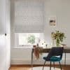 Grey geometric diamond-patterned Roman blind, lowered over the window and filtering soft daylight; small home office desk beneath with teal chair, vase of flowers, makeup mirror and framed coastal print.