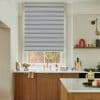 Gray cellular roller blind with horizontal striped bands, lowered to cover most of the window and filtering daylight, above a kitchen sink with brass faucet, marble countertop and wooden cabinets.