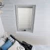 White pleated blind, fully lowered in a skylight recess with fine horizontal folds; diffusing daylight in a white-painted slatted attic bedroom above a bed with pillows and a guitar cushion.