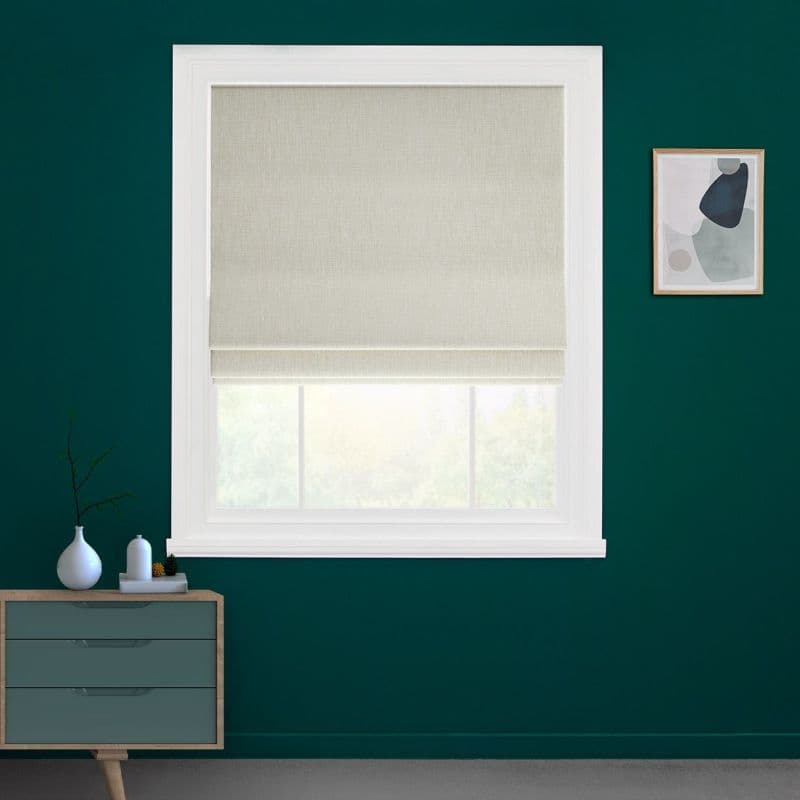 Beige fabric Roman blind, lowered with a small bottom fold across a white-framed window; filtering soft daylight; set on a teal-painted wall above a mid-century dresser and framed abstract art.
