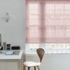 Sheer pink Roman blind, lowered over a large multi-pane sash window, softly filtering daylight. Context: minimalist home office with white desk, wooden chair, paper pendant lamp and leafless trees outside.