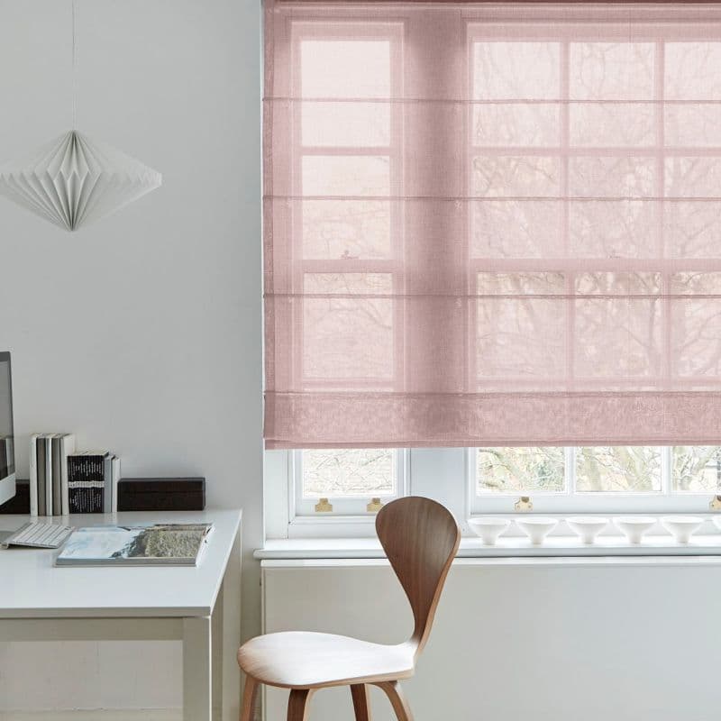 Sheer pink Roman blind, lowered over a large multi-pane sash window, softly filtering daylight. Context: minimalist home office with white desk, wooden chair, paper pendant lamp and leafless trees outside.