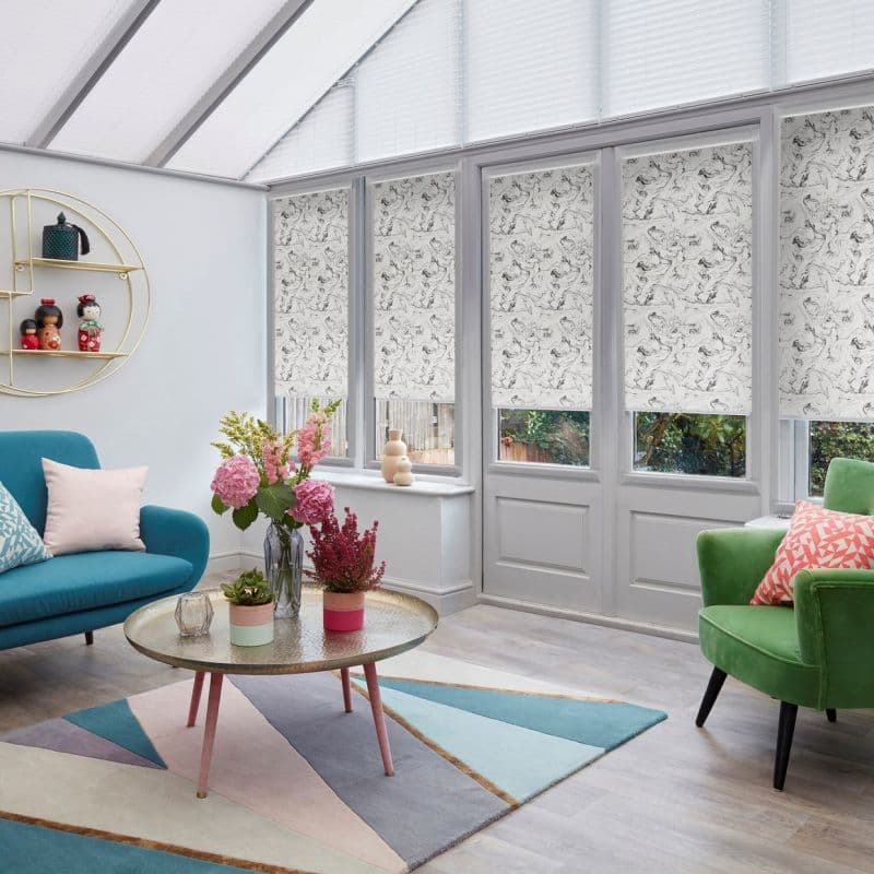 Marbled black-and-white roller blinds covering tall French windows and doors, lowered to mid-height and filtering light; bright conservatory-style living room with skylight, colorful sofas, rug, and plants.