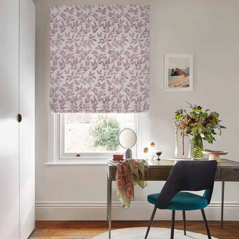 A floral-patterned Roman blind, lowered and partially covering the window, patterned with purple sprigs; filtering soft daylight into a tidy home office with desk, chair, and a vase of flowers.