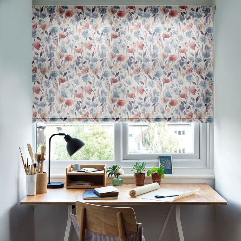 Floral roller blind lowered across the window, watercolor blue‑and‑pink flowers; filtering soft daylight into a small home workspace — wooden desk with lamp, art brushes, notebooks and potted plants.