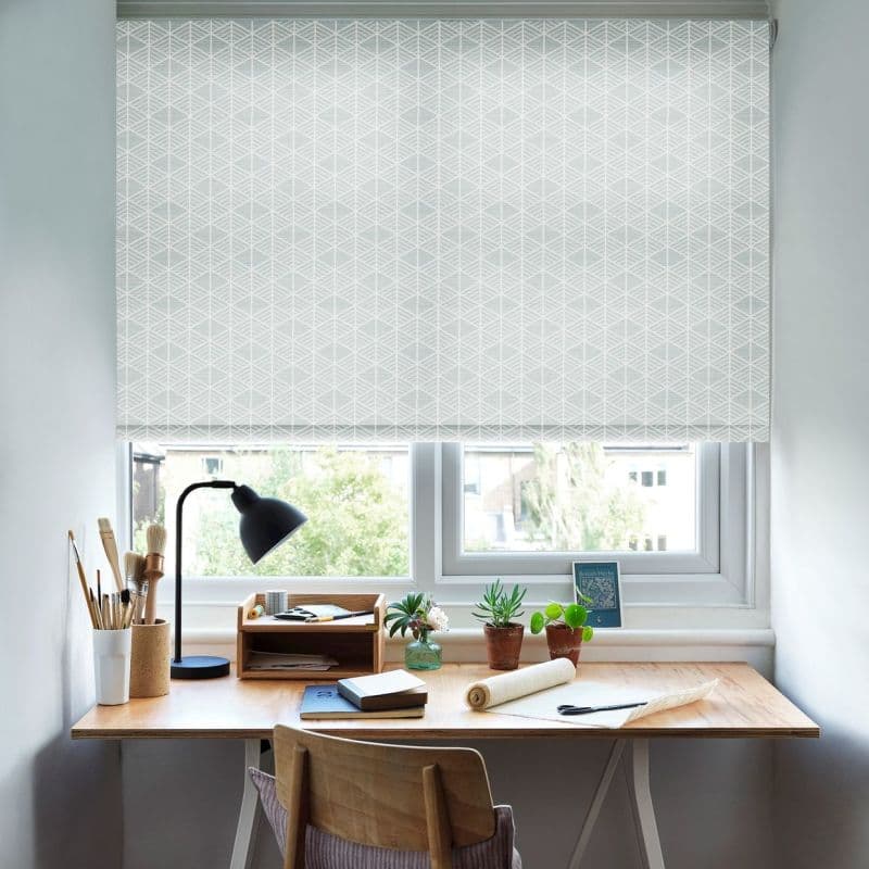 White geometric roller blind lowered and diffusing daylight; below, a wooden desk with lamp, art brushes, notebooks, a rolled fabric, potted succulents, and a view of trees and nearby buildings.