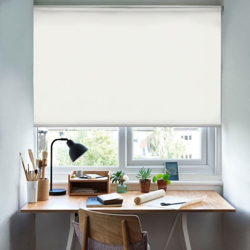 White roller blind lowered across a double window, blocking direct view and diffusing daylight; above a wooden desk with lamp, brushes, small potted plants and chair in a bright workspace.