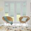 Pleated cellular blinds (seafoam with horizontal texture) covering French doors and sidelights, lowered mid‑height and filtering daylight; modern bright sitting area with two rattan chairs, patterned rug, lawn view.
