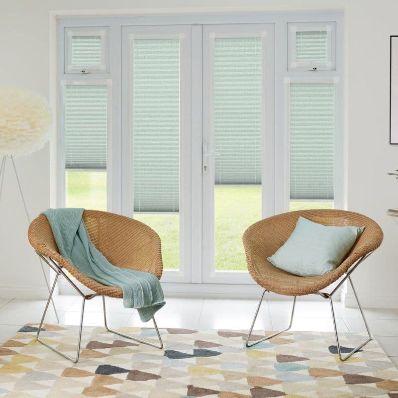 Pleated cellular blinds (seafoam with horizontal texture) covering French doors and sidelights, lowered mid‑height and filtering daylight; modern bright sitting area with two rattan chairs, patterned rug, lawn view.