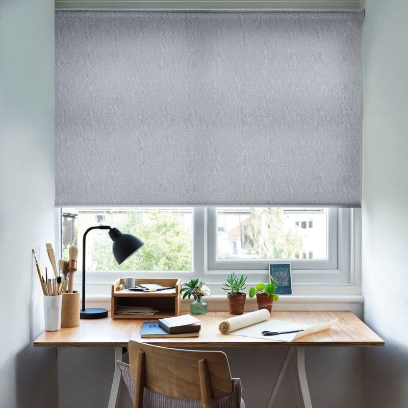 Light-gray textured roller blind lowered across a double window, filtering daylight and obscuring most view; below, tidy wooden desk with lamp, brushes, notebooks and potted plants in bright workspace.