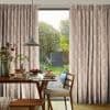Pleated curtains (pale pink with white feather pattern) framing a sliding glass door, slightly parted and filtering daylight; dining table set for breakfast, pendant lamp, view of a green garden.