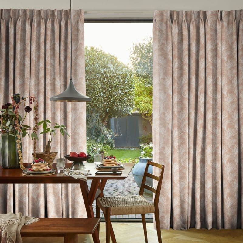 Pleated curtains (pale pink with white feather pattern) framing a sliding glass door, slightly parted and filtering daylight; dining table set for breakfast, pendant lamp, view of a green garden.