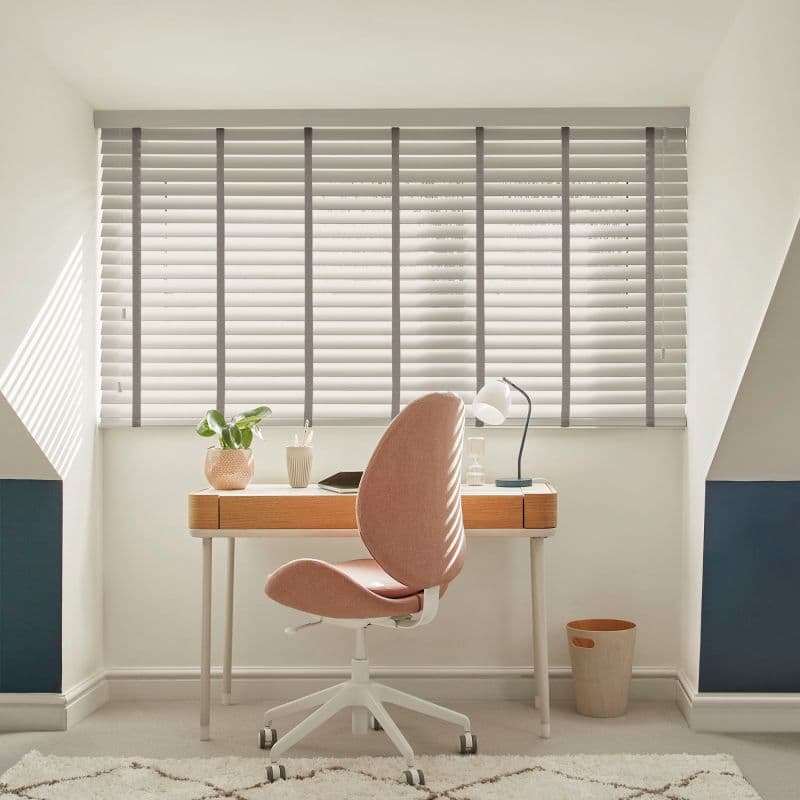 Horizontal slatted blinds over a wide window, pale grey with tapes and cord, closed and filtering daylight, casting striped shadows across a study with wooden desk and pink swivel chair.