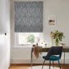 Gray patterned Roman blind lowered over window, filtering daylight; wooden desk with round mirror, vase of flowers and scarf beside a teal chair in a lit office with garden view.