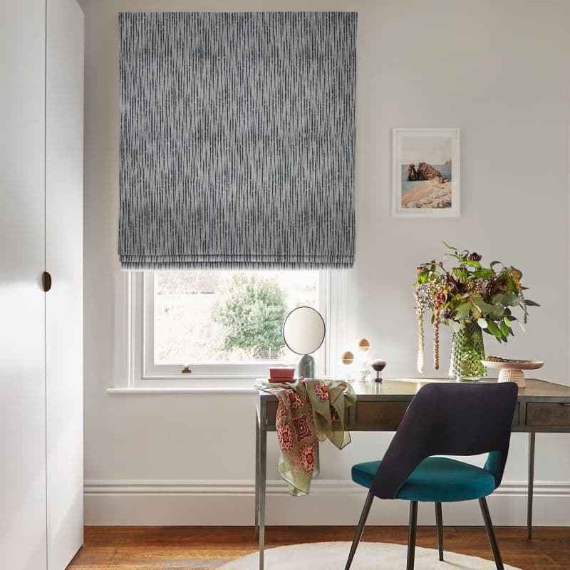 Gray patterned Roman blind lowered over window, filtering daylight; wooden desk with round mirror, vase of flowers and scarf beside a teal chair in a lit office with garden view.