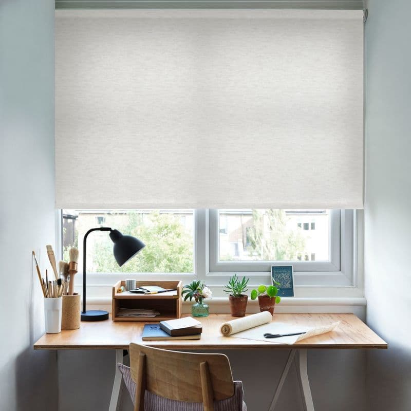 White roller blind, lowered across a double window, diffusing daylight; a wooden desk beneath holds a black desk lamp, potted succulents, brushes and notebooks in a compact, well-lit home workspace.