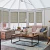Pleated cellular blinds covering five tall bay windows, lowered to mid-height and filtering soft daylight; bright conservatory-style living room with beige sofa, wooden coffee table and pastel accents.