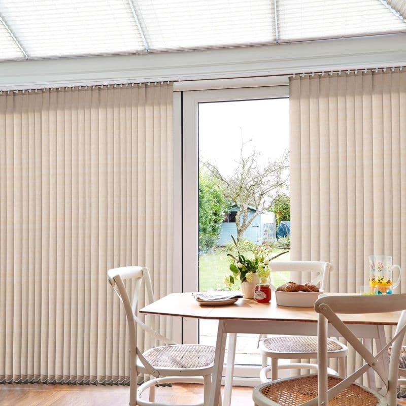 Beige vertical blinds, full-height slatted panels drawn closed at sides and parted at center, partially opening and filtering daylight through a glass patio door into a bright dining area.