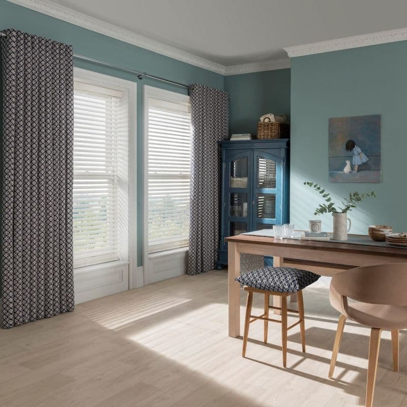 Patterned curtains and white horizontal blinds at tall windows, blinds filtering sunlight through slats — casting striped light across pale wood floor in teal-walled dining room with table and blue cabinet.