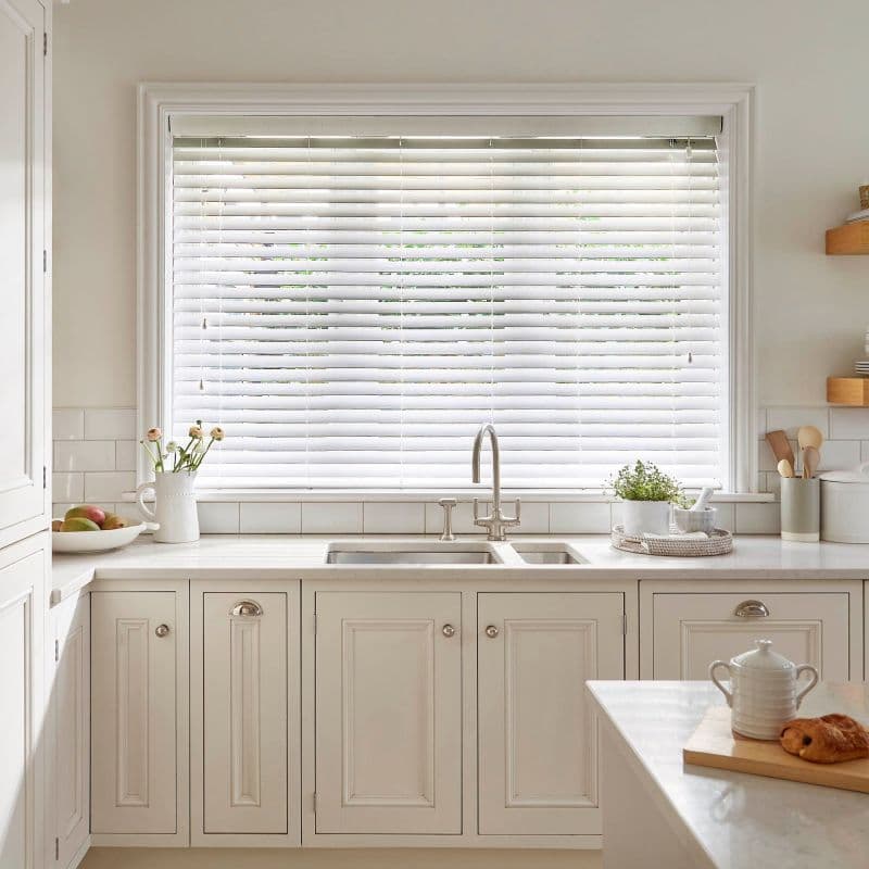 White horizontal blinds lowered and slightly tilted, filtering daylight over a chrome faucet and double sink in a bright neutral kitchen with marble counters, paneled cabinets, and potted herbs.