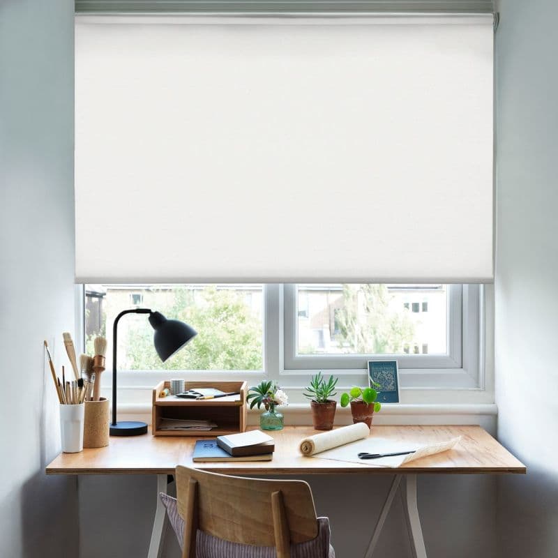 White roller blind lowered, diffusing daylight; wooden desk beneath holds a black lamp, paintbrushes, notebooks, small potted plants, rolled canvas and chair in a bright home workspace with outside greenery.