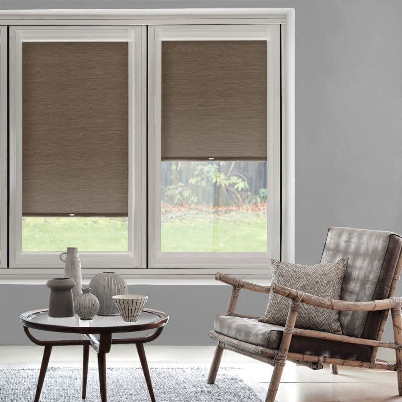 Brown fabric roller shades, lowered across two adjacent windows (left fully, right partially), filtering daylight; neutral living room with wooden-framed leather armchair, patterned cushion and low coffee table with ceramics.