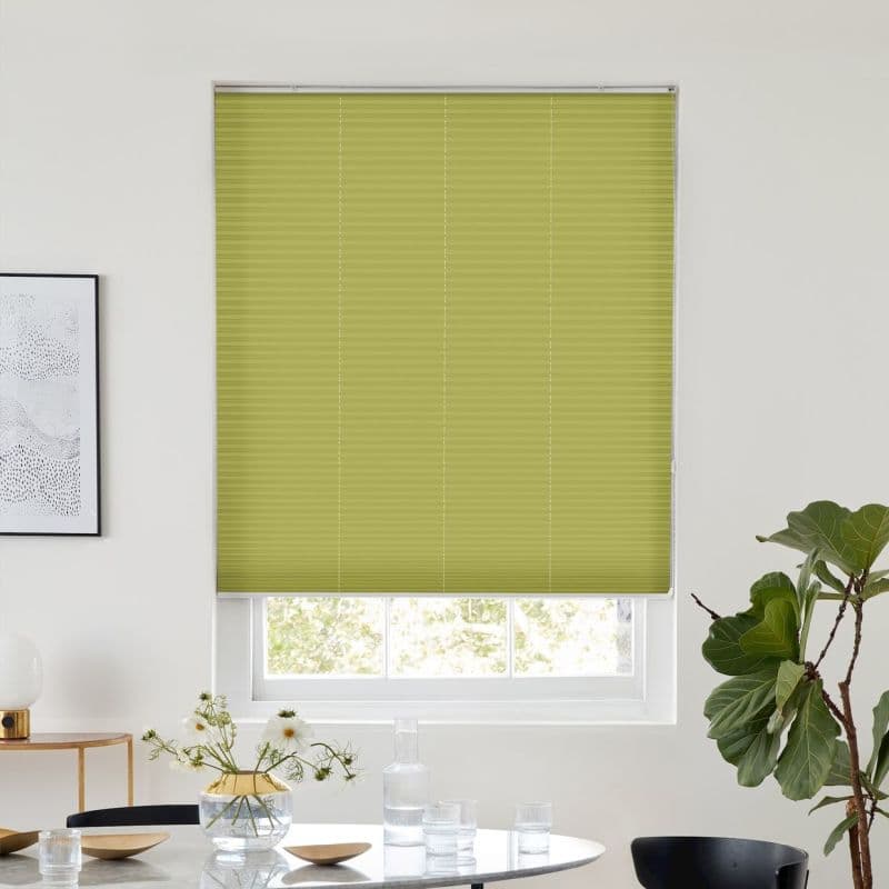 Green pleated cellular blind, fully lowered across the window, filtering light through a narrow bottom gap; above a dining table with glassware, framed art, and a potted fiddle‑leaf fig.