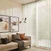 Sheer cream vertical blinds covering a full-height window, filtering sunlight into soft striped shadows across a living room with a beige sofa, textured cushions and framed wall art.