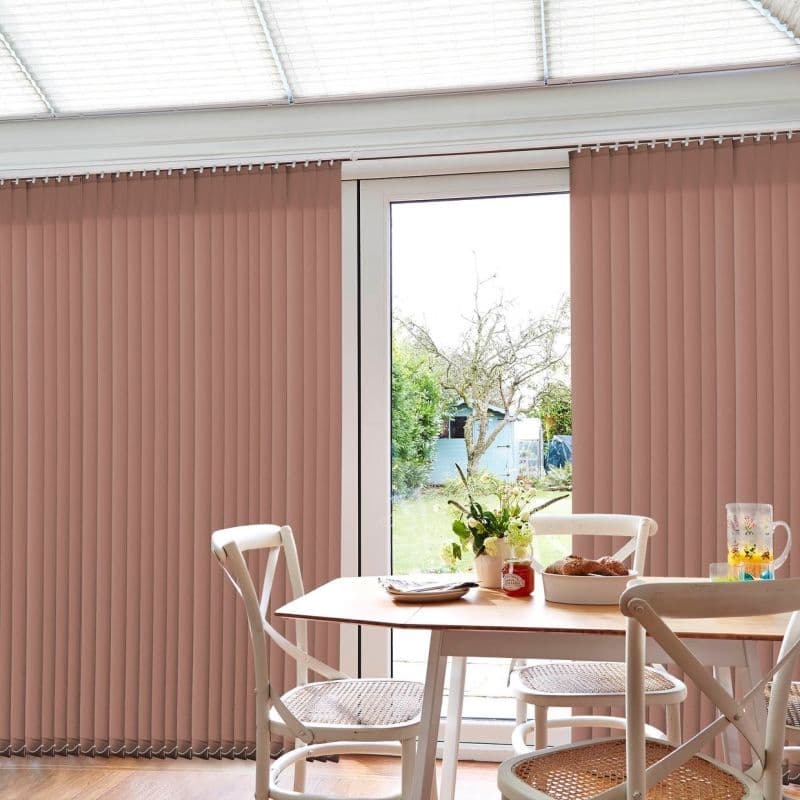 Vertical rose-beige vertical blinds covering a patio sliding glass door, mostly closed with a central gap; filtering daylight into a sunlit dining area with table, chairs, flowers and garden view.