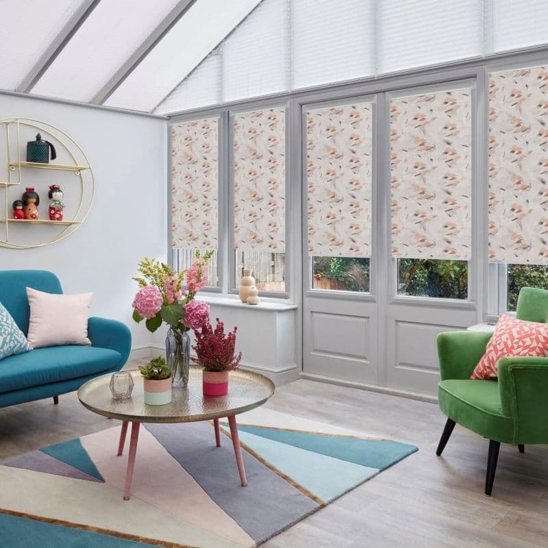Patterned roller blinds (soft beige/peach leaf print) covering tall French doors and windows, partially lowered and filtering daylight; in a bright conservatory-style living room with pastel furnishings and plants.