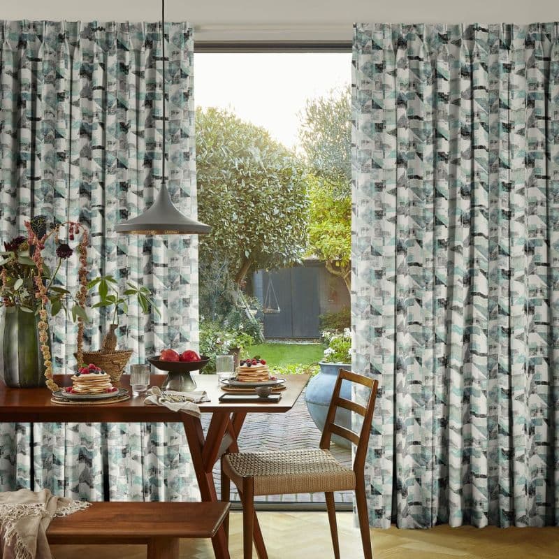 Pleated teal-gray geometric curtains, full-length, frame a central open sliding door, parted to admit sunlight; a dining table set for breakfast overlooks a garden with trees, swing, and shed.