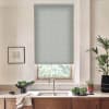A patterned roller blind covers a kitchen window above a stainless-steel sink and faucet, set in a wooden-cabinet countertop with potted plants, a vegetable bowl, and framed art.