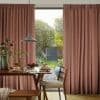 Thick pleated dusty-rose curtains drawn across a large sliding glass door, filtering soft daylight into a dining nook with a wooden table set for breakfast and a green garden visible outside.