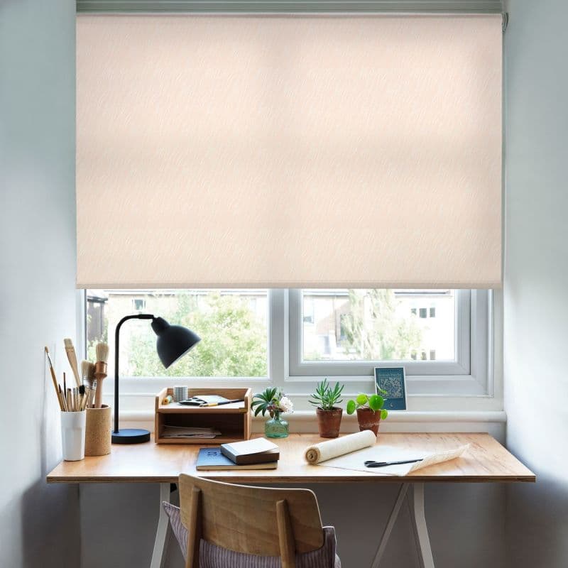 A beige patterned roller blind, lowered fully with subtle wavy texture, softly diffusing daylight; above a wooden desk in a bright home office/studio with a lamp, plants, brushes and notebooks.