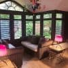 Dark wooden plantation shutters cover a bay of tall windows, slats partially open, filtering garden light into a cozy sunroom with wicker sofas, neutral cushions, wooden floor and colorful chandelier.