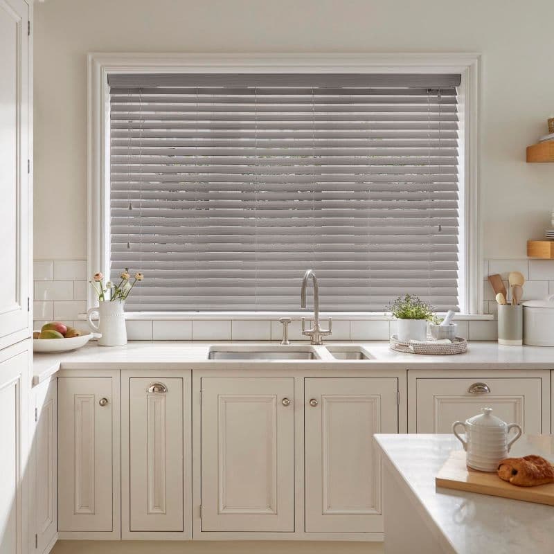 Gray horizontal Venetian blinds, fully lowered and closed, slightly filtering daylight; in a bright cream kitchen over a double sink with marble countertop, faucet, plants, and countertop accessories.
