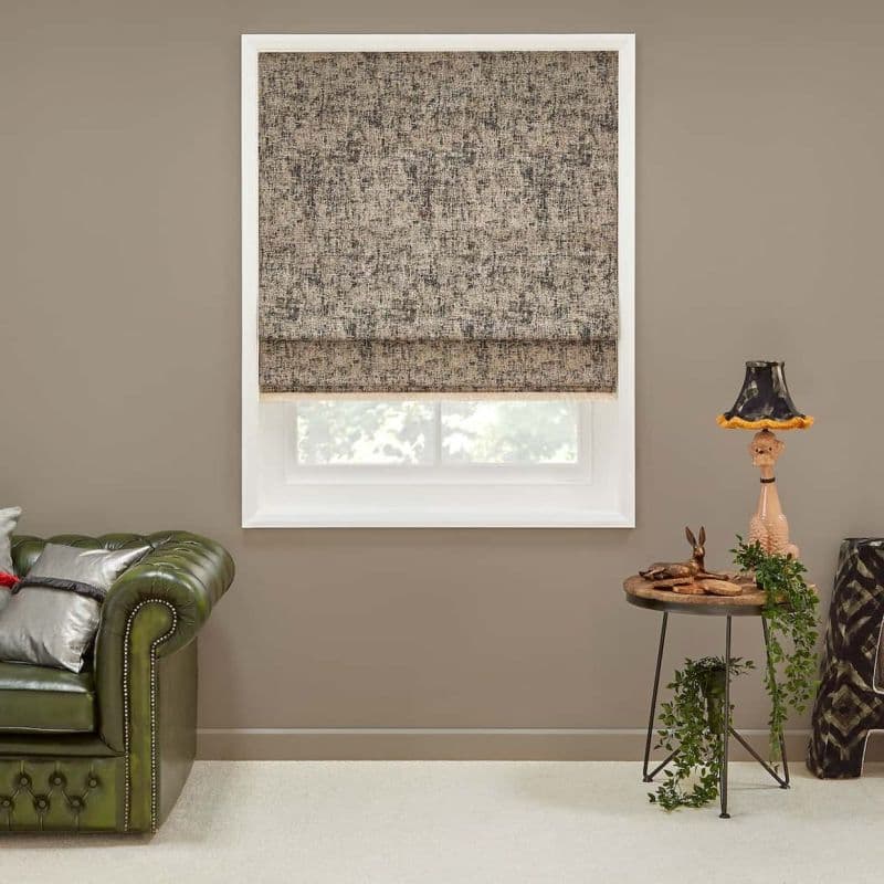 Textured beige-gray Roman shade, mostly lowered with visible hem, filtering daylight through a white-framed window; set in a neutral living room with green leather sofa, side table, lamp, and trailing plant.