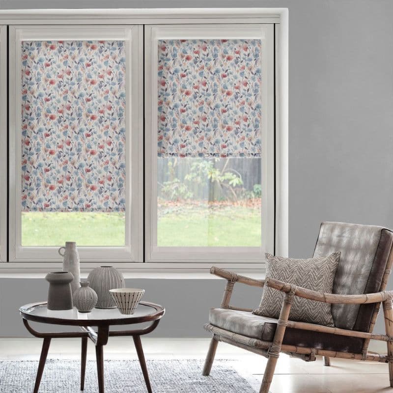 Floral-patterned roller blinds covering twin sash windows—left fully lowered, right partially lowered—filter soft daylight. In a gray living room with a rattan armchair, cushion, round coffee table and garden view.