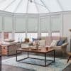 Pale honeycomb cellular blinds lowered across tall conservatory windows, closed and softly filtering daylight; cozy sitting area with beige sofa, wooden coffee table, blue rug and pastel cushions.