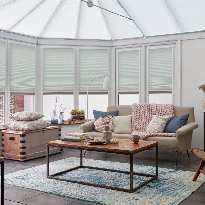 Pale honeycomb cellular blinds lowered across tall conservatory windows, closed and softly filtering daylight; cozy sitting area with beige sofa, wooden coffee table, blue rug and pastel cushions.