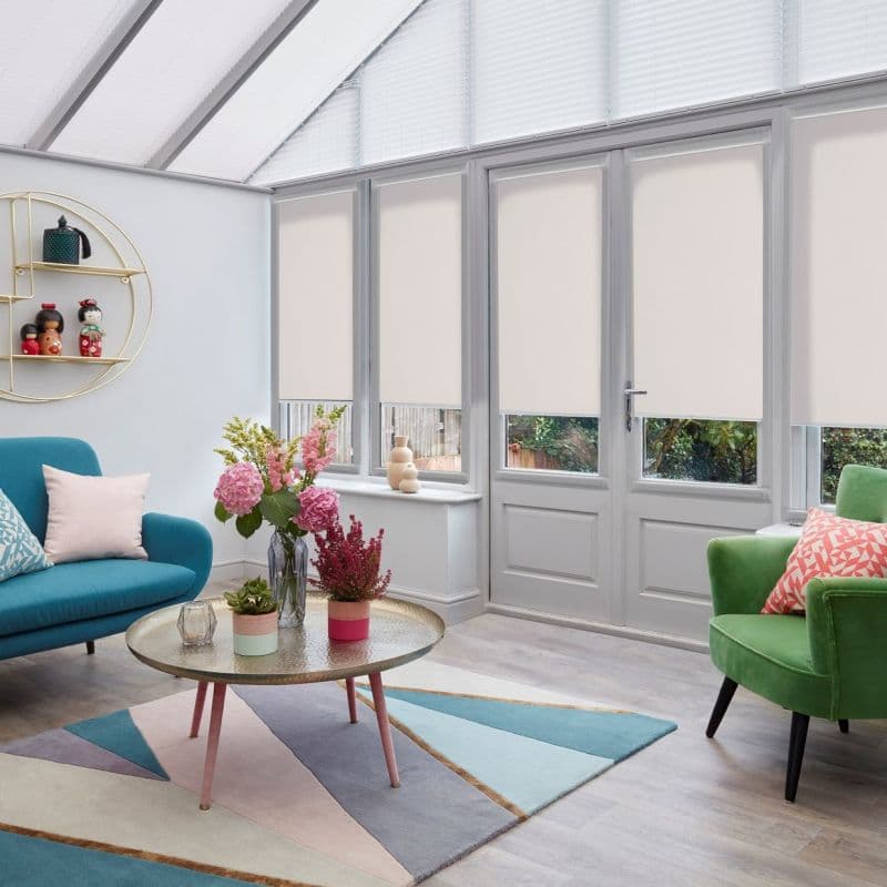 Beige roller blinds lowered to mid-height across tall conservatory windows and glass door, filtering light; bright modern lounge with colorful sofas, floral centerpiece, and glimpses of a garden.